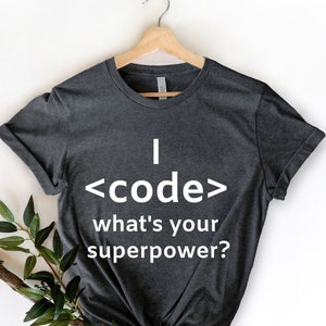 May include: Dark gray t-shirt with the text "I code what's your superpower?" in white. The shirt is hanging on a wooden hanger against a white background. A sprig of greenery is in the lower left corner.