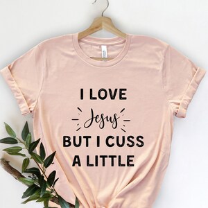 May include: A pale pink t-shirt with the text "I LOVE Jesus BUT I CUSS A LITTLE" in black. The shirt is displayed on a wooden hanger. A sprig of greenery is in the lower left corner, against a white backdrop.