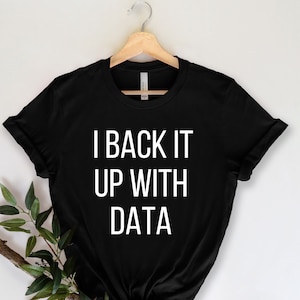 Könnte beinhalten: Schwarzes T-Shirt mit dem weißen Text "I BACK IT UP WITH DATA". Das Shirt hängt an einem Holzbügel. Ein Zweig mit Grünzeug befindet sich links vom Shirt.