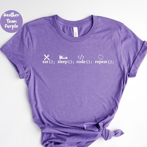 May include: Purple t-shirt with a white graphic of a fork and knife, a bed, code symbols, and a refresh icon. The text below the icons reads: "eat(); sleep(); code(); repeat();"