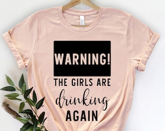 Pode incluir: Uma t-shirt rosa claro com o texto preto "WARNING! THE GIRLS ARE drinking AGAIN". A t-shirt está pendurada num cabide de madeira. Um ramo de vegetação é colocado no lado inferior esquerdo da t-shirt.