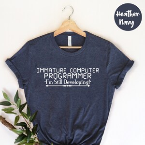 Op de afbeelding: Heather navy t-shirt met de witte tekst "IMMATURE COMPUTER PROGRAMMER - I'm Still Developing!". Het shirt hangt aan een houten hanger met een takje groen.
