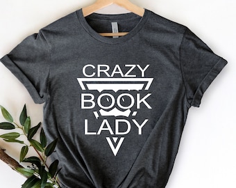 Può includere: T-shirt grigio scuro con la scritta bianca "CRAZY BOOK LADY" in un design triangolare. La maglietta è appesa a una gruccia di legno su sfondo bianco. Un rametto di verde è in basso a sinistra.
