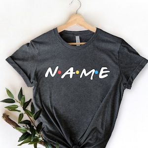 May include: A dark grey t-shirt with the word "NAME" in white letters with colorful dots between each letter. The shirt is hanging on a wooden hanger.