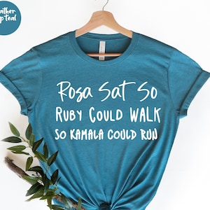 May include: A teal blue t-shirt with white text that reads "Rosa Sat So Ruby Could Walk So Kamala Could Run".