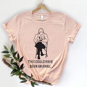 Könnte beinhalten: Hellrosa T-Shirt mit einer schwarzen Grafik einer Person, die auf einem Stuhl sitzt und eine Brille und Fäustlinge trägt. Der Text "THIS COULD HAVE BEEN AN EMAIL" ist darunter gedruckt.