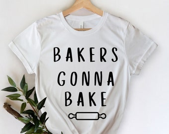 Peut inclure: T-shirt blanc avec l'inscription noire "BAKERS GONNA BAKE" et un dessin de rouleau à pâtisserie. Le t-shirt est suspendu à un cintre en bois, avec une branche et des feuilles vertes sur le côté.