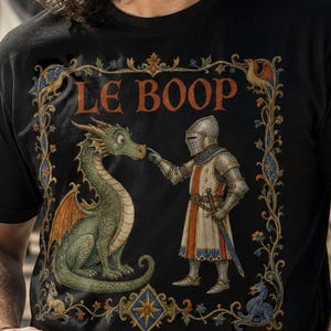 May include: A black t-shirt featuring a graphic print of a knight in armour touching the nose of a green dragon. The design is enclosed in an ornate, medieval-style rectangular border with the text LE BOOP printed in a serif font at the top centre.