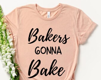 Peut inclure: T-shirt pêche clair avec l'inscription noire "Bakers Gonna Bake". Le t-shirt est noué en bas et est orné d'un bouquet de fleurs blanches avec des tiges vertes.