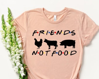 Könnte beinhalten: Hellrosa T-Shirt mit dem schwarzen Schriftzug "FRIENDS NOT FOOD", im Stil der Fernsehserie Friends. Das Design zeigt schwarze Silhouetten eines Huhns, einer Kuh und eines Schweins, für eine vegane Botschaft.