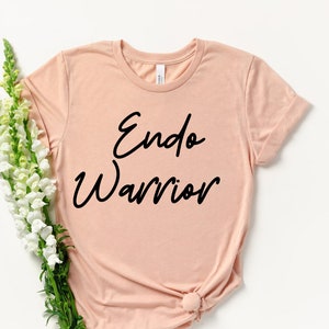 May include: A light peach-colored t-shirt with the words "Endo Warrior" in black script. The shirt is styled with a knot at the bottom and is accompanied by a bouquet of white and green flowers. The t-shirt is made of soft material.