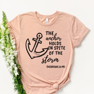 May include: A pale peach t-shirt featuring a black anchor design and the words "The anchor holds in spite of the storm (Hebrews 6:19)". The shirt is knotted at the hem and styled with a bouquet of white flowers.