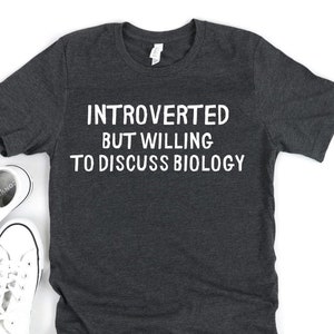 May include: Dark gray t-shirt with the text "INTROVERTED BUT WILLING TO DISCUSS BIOLOGY" in white. Two pairs of white canvas sneakers are next to the shirt on a white surface.