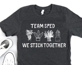 Pode incluir: Camiseta cinza escuro com o texto branco "TEAM SPED WE STICK TOGETHER". Apresenta quatro ilustrações de plantas. Um par de tênis de lona brancos com acabamento preto está ao lado.