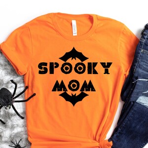 May include: An orange t-shirt with the words "SPOOKY MOM" in black block letters, with bat and circle designs. The shirt is paired with dark blue jeans. A grey spider web and black spiders are in the background.