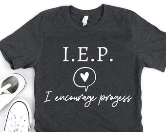 Pode incluir: Camiseta cinza escuro com o texto "I.E.P. I encourage progress" em branco. Um coração branco está dentro de um balão de fala. Dois pares de tênis brancos estão no lado esquerdo da camiseta.