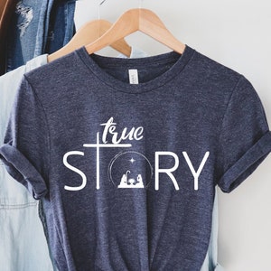 May include: A navy blue t-shirt with the words "true STORY" in white. The word "true" is above a white cross. The word "STORY" is above a circular design with a star and a nativity scene. The shirt is hanging on a wooden hanger.