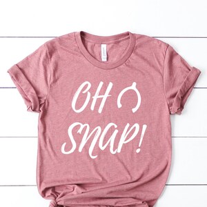 May include: A dusky pink t-shirt with the words "OH SNAP!" in white script. The sleeves are folded and the bottom is knotted. The background is a white wooden surface.