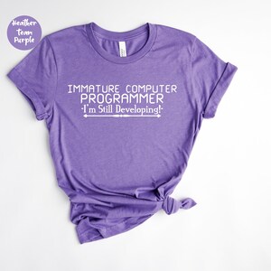 May include: A heather purple t-shirt featuring the white text: "IMMATURE COMPUTER PROGRAMMER - I'm Still Developing!". The shirt has a crew neck and rolled sleeves. The fabric appears soft and comfortable.
