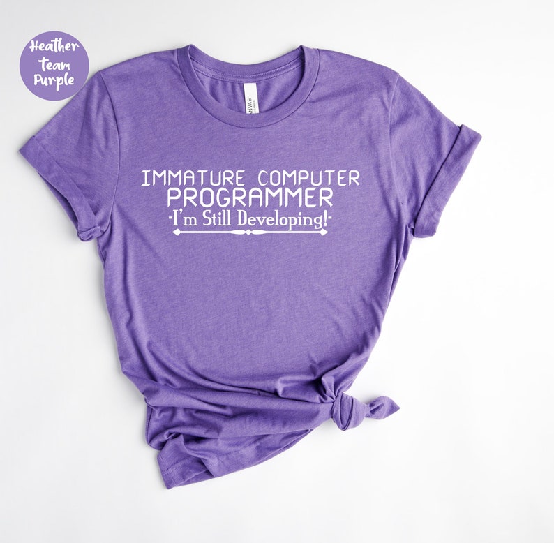 May include: A heather purple t-shirt featuring the white text: "IMMATURE COMPUTER PROGRAMMER - I'm Still Developing!". The shirt has a crew neck and rolled sleeves. The fabric appears soft and comfortable.