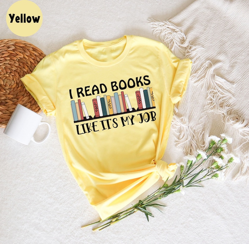 May include: A yellow t-shirt with the text "I READ BOOKS LIKE IT'S MY JOB" in black. The design features a row of colourful books. The shirt is on a white surface with a woven basket and a white mug in the background.
