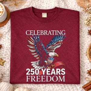 May include: Maroon t-shirt with a graphic of an eagle with red, white, and blue wings, holding a shield and ribbon. The text reads "CELEBRATING 250 YEARS FREEDOM". The shirt is displayed on a white, textured surface.