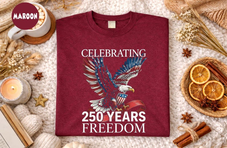 May include: Maroon t-shirt with a graphic of an eagle with red, white, and blue wings, holding a shield and ribbon. The text reads "CELEBRATING 250 YEARS FREEDOM". The shirt is displayed on a white, textured surface.