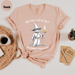 May include: Peach-coloured t-shirt with a cartoon wizard frog holding a fireball. The text reads "YOU TALK A LOT OF SHIT FOR SOMEONE IN FIREBALL RANGE." The shirt is displayed on a white surface with a coffee cup and notepad.