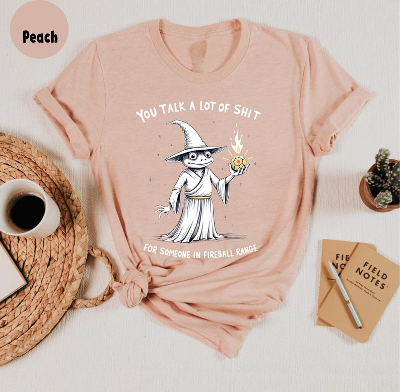 May include: Peach-coloured t-shirt with a cartoon wizard frog holding a fireball. The text reads "YOU TALK A LOT OF SHIT FOR SOMEONE IN FIREBALL RANGE." The shirt is displayed on a white surface with a coffee cup and notepad.