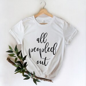 May include: White t-shirt with the phrase "all peopled out" in black script. The shirt is hanging on a wooden hanger, with a sprig of green leaves and a branch in the lower left corner. The shirt is made of a soft material.