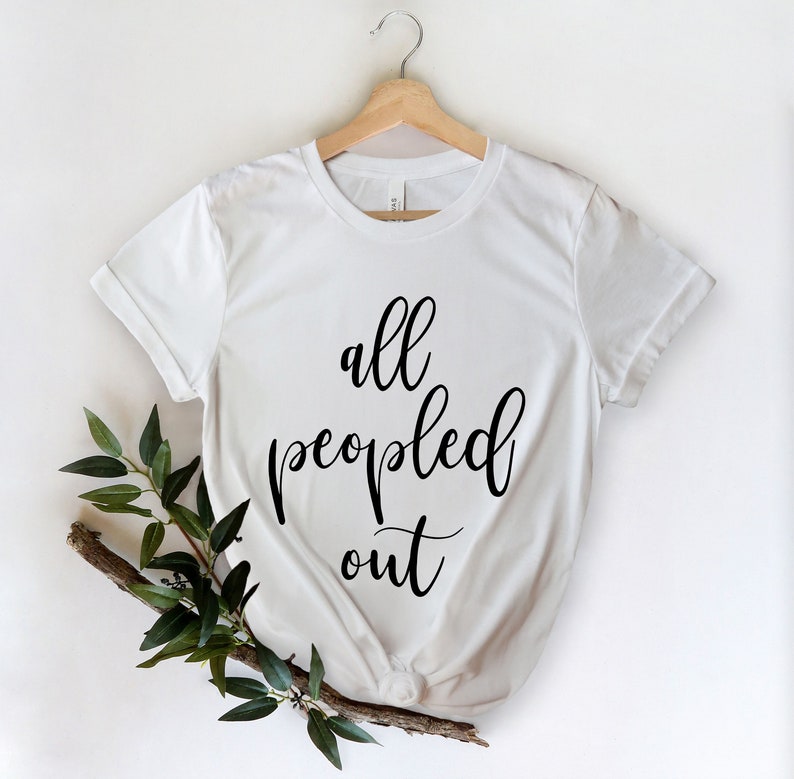 May include: White t-shirt with the phrase "all peopled out" in black script. The shirt is hanging on a wooden hanger, with a sprig of green leaves and a branch in the lower left corner. The shirt is made of a soft material.