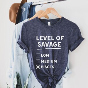 Op de afbeelding: Een marineblauw t-shirt met de witte tekst "LEVEL OF SAVAGE". Het shirt heeft een afbeelding met selectievakjes voor "LOW", "MEDIUM" en "PISCES". Het shirt hangt aan een houten hanger met andere kledingstukken.