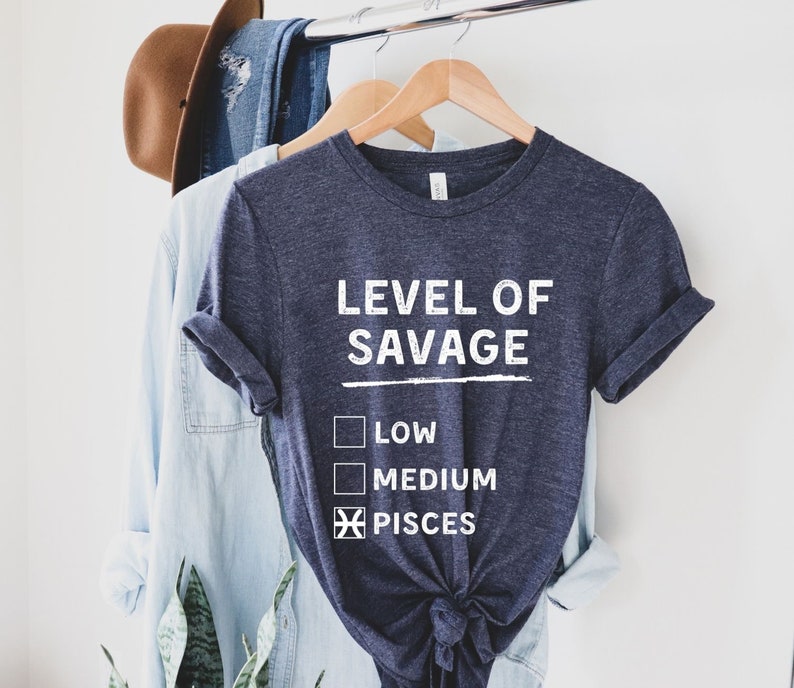 Op de afbeelding: Een marineblauw t-shirt met de witte tekst "LEVEL OF SAVAGE". Het shirt heeft een afbeelding met selectievakjes voor "LOW", "MEDIUM" en "PISCES". Het shirt hangt aan een houten hanger met andere kledingstukken.