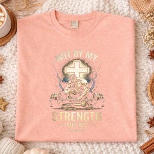 K&ouml;nnte beinhalten: Hellpfirsichfarbenes T-Shirt mit einem grafischen Design. Das Design zeigt ein Kreuz, einen Gewichtheber und den Text "NOT BY MY STRENGTH BUT HIS LOVE". Das Shirt wird auf einer wei&szlig;en, strukturierten Oberfl&auml;che pr&auml;sentiert.