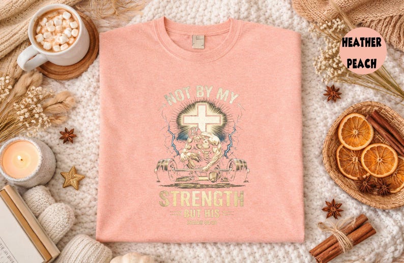 K&ouml;nnte beinhalten: Hellpfirsichfarbenes T-Shirt mit einem grafischen Design. Das Design zeigt ein Kreuz, einen Gewichtheber und den Text "NOT BY MY STRENGTH BUT HIS LOVE". Das Shirt wird auf einer wei&szlig;en, strukturierten Oberfl&auml;che pr&auml;sentiert.