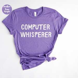 May include: A heather purple t-shirt with the words "COMPUTER WHISPERER" printed in white. The shirt is knotted at the bottom. A small circular graphic in the upper left corner reads "Heather Team Purple".