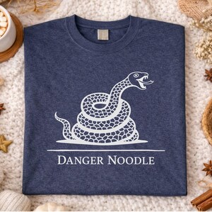Peut inclure: T-shirt bleu marine avec un motif blanc d'un serpent enroul&eacute; et la mention "DANGER NOODLE" en dessous. L'image est pr&eacute;sent&eacute;e sur une surface textur&eacute;e et douce, avec des &eacute;l&eacute;ments d&eacute;coratifs.