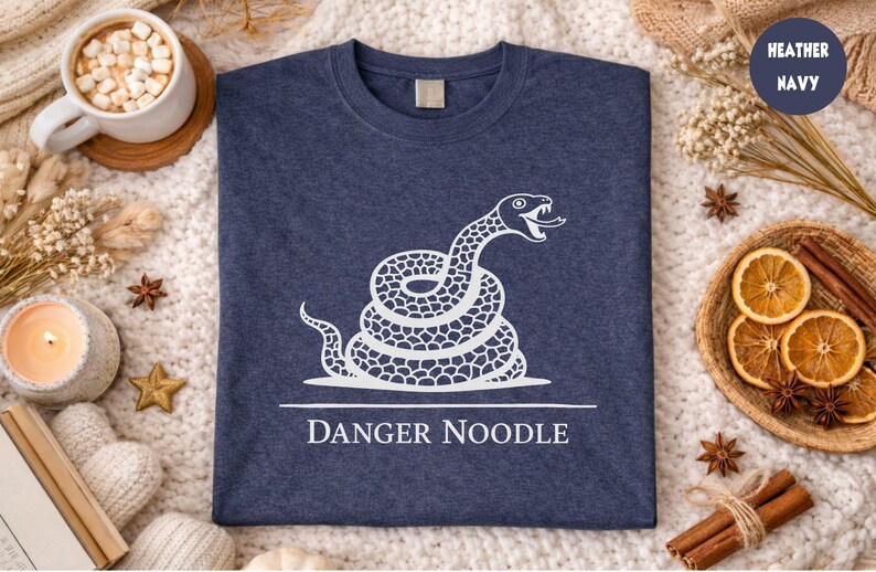 Peut inclure: T-shirt bleu marine avec un motif blanc d'un serpent enroul&eacute; et la mention "DANGER NOODLE" en dessous. L'image est pr&eacute;sent&eacute;e sur une surface textur&eacute;e et douce, avec des &eacute;l&eacute;ments d&eacute;coratifs.