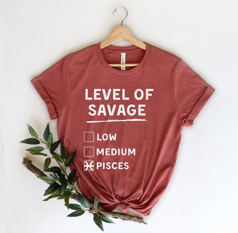 Op de afbeelding: Een roestkleurig T-shirt met de witte tekst "LEVEL OF SAVAGE". Daaronder staan selectievakjes met "LOW", "MEDIUM" en "PISCES", waarbij het vakje "PISCES" is aangevinkt. Het shirt hangt aan een houten hanger.