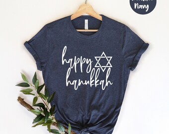 Könnte beinhalten: Marineblaues T-Shirt mit dem weißen Schriftzug "happy hanukkah" und einem weißen Davidstern. Das T-Shirt hängt an einem Holzbügel, mit einem Zweig Grünzeug an der Seite.