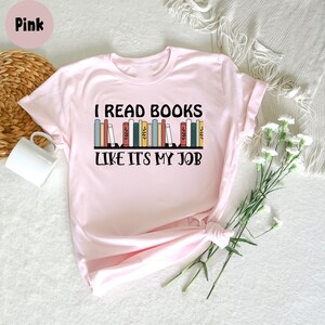May include: A pale pink t-shirt featuring the text "I READ BOOKS LIKE IT'S MY JOB" in black, with a design of colourful books. The shirt is folded, with a white mug and flowers in the background.