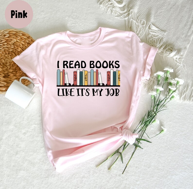 May include: A pale pink t-shirt featuring the text "I READ BOOKS LIKE IT'S MY JOB" in black, with a design of colourful books. The shirt is folded, with a white mug and flowers in the background.