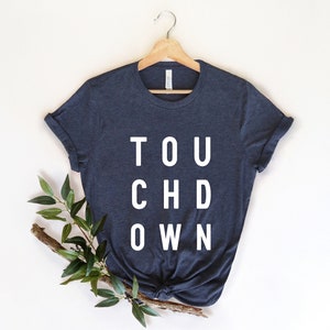 Könnte beinhalten: Marineblaues T-Shirt mit dem weißen Aufdruck "TOUCHDOWN", aufgehängt an einem Holzbügel. Die Ärmel sind hochgekrempelt, und das Shirt ist mit einem Zweig mit grünen Blättern verziert.