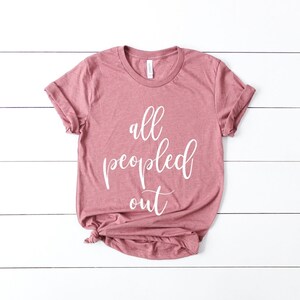 May include: A dusty rose-colored t-shirt with the phrase "all peopled out" in white script. The shirt is folded at the bottom and has rolled-up sleeves. The t-shirt is laying on a white wooden surface.
