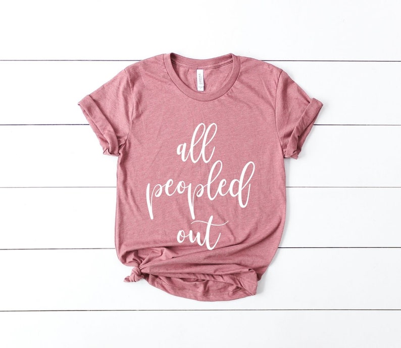 May include: A dusty rose-colored t-shirt with the phrase "all peopled out" in white script. The shirt is folded at the bottom and has rolled-up sleeves. The t-shirt is laying on a white wooden surface.