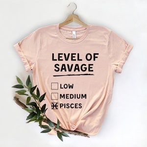 Op de afbeelding: Een lichtroze t-shirt met de zwarte tekst "LEVEL OF SAVAGE". Daaronder staan drie selectievakjes, met "PISCES" aangevinkt. Het t-shirt hangt aan een houten hanger.