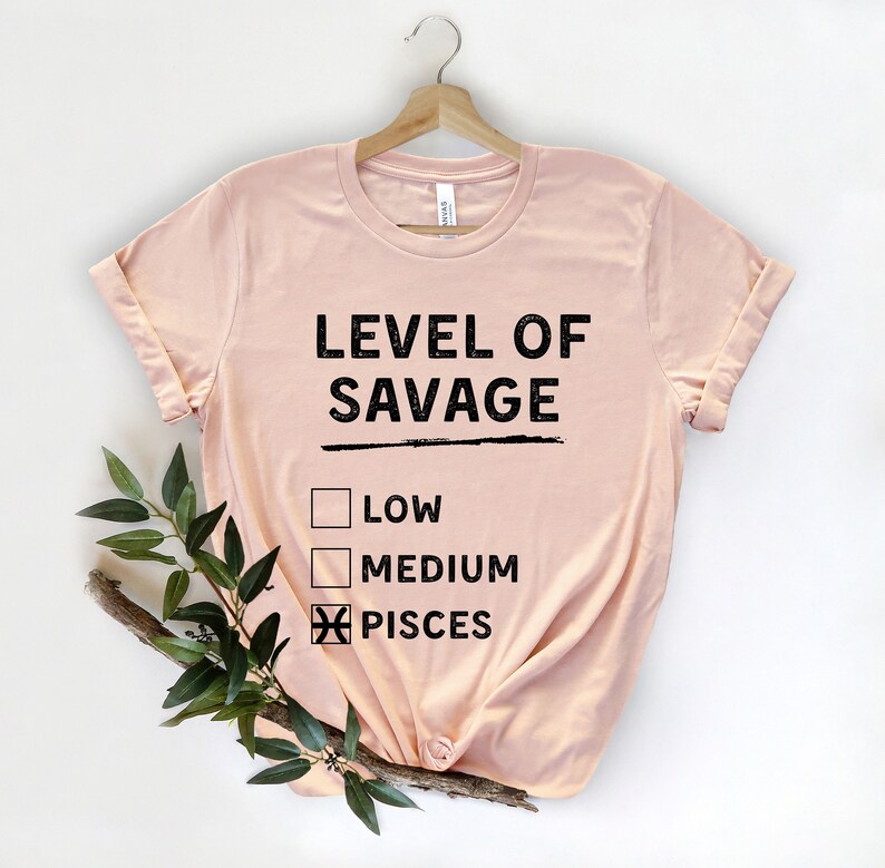 Op de afbeelding: Een lichtroze t-shirt met de zwarte tekst "LEVEL OF SAVAGE". Daaronder staan drie selectievakjes, met "PISCES" aangevinkt. Het t-shirt hangt aan een houten hanger.