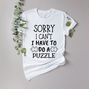 Pode incluir: Camiseta branca com o texto preto "SORRY I CAN'T I HAVE TO DO A PUZZLE". O design inclui um gr&aacute;fico de pe&ccedil;a de quebra-cabe&ccedil;a. A camiseta &eacute; exibida em um fundo neutro com folhagem verde.