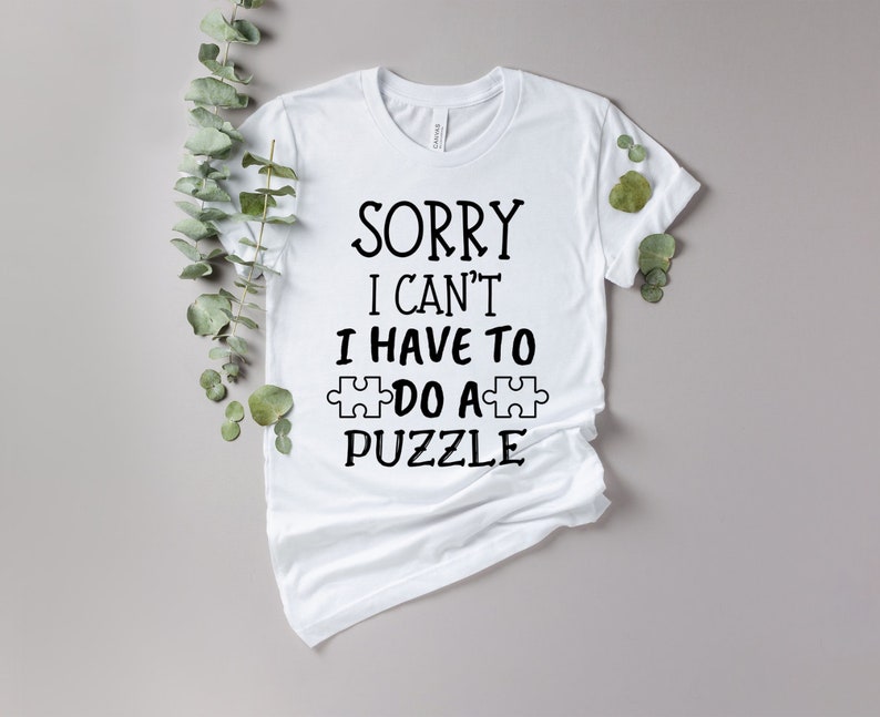 Pode incluir: Camiseta branca com o texto preto "SORRY I CAN'T I HAVE TO DO A PUZZLE". O design inclui um gr&aacute;fico de pe&ccedil;a de quebra-cabe&ccedil;a. A camiseta &eacute; exibida em um fundo neutro com folhagem verde.