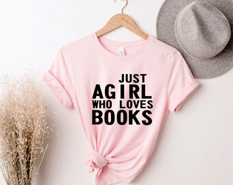 Può includere: Una maglietta rosa chiaro con la scritta nera "JUST A GIRL WHO LOVES BOOKS". La maglietta è appesa a una gruccia di legno. Un cappello grigio è visibile sullo sfondo, contro un muro bianco.